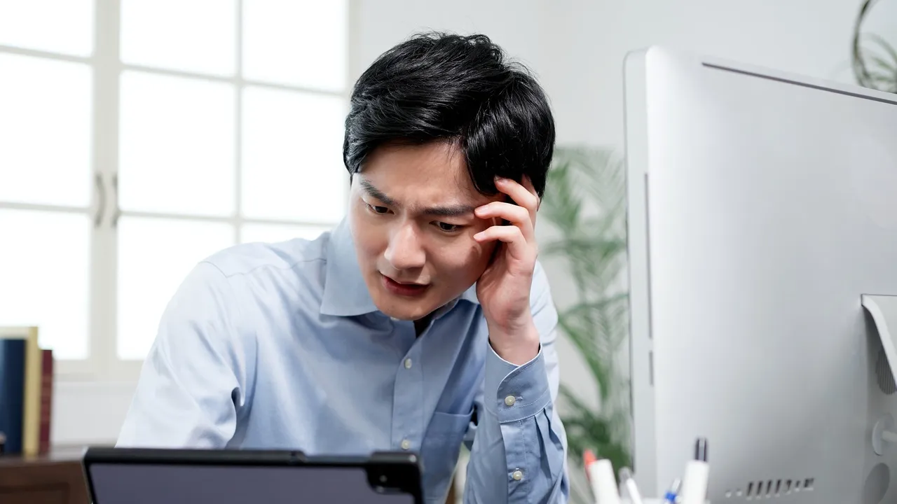 Concerned man in a blue shirt sits at a desk, looking at a tablet with a large computer monitor nearby in a bright office