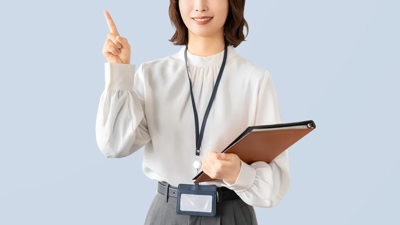 Professional woman in a white blouse points upward while holding a brown notebook and a clipboard against a light blue background, wearing a lanyard with a badge.