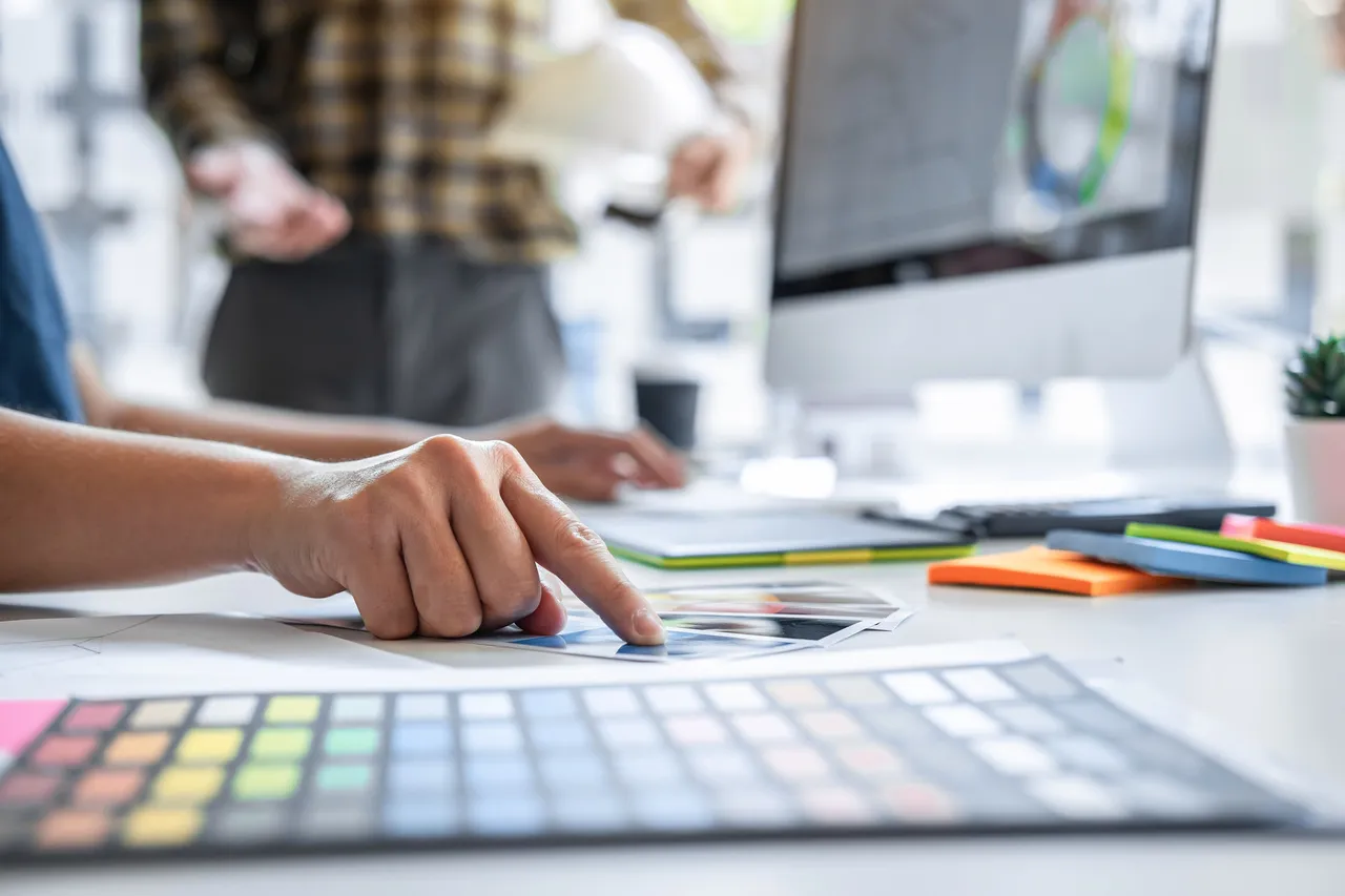 Designer at a bright desk compares color swatches and printed images with a computer monitor in the background.