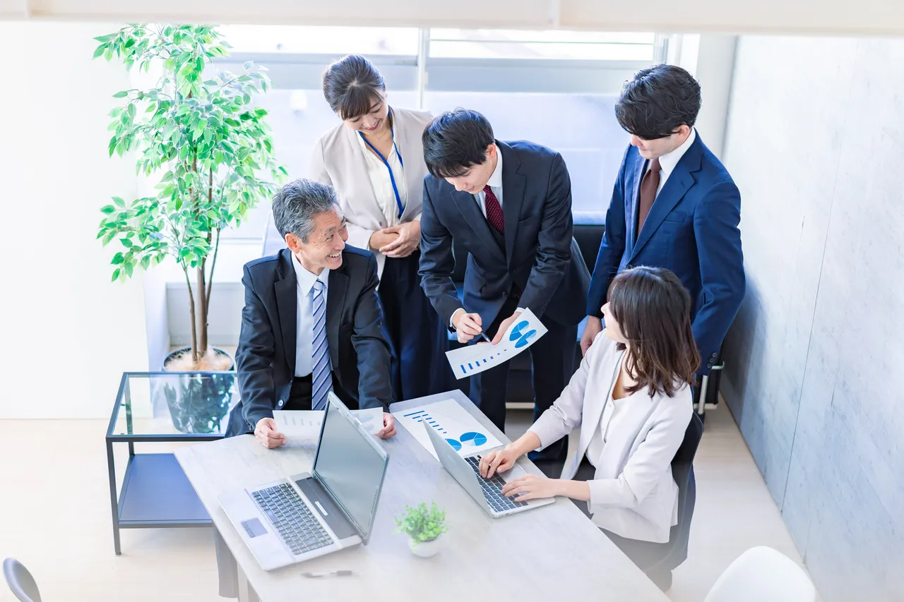 Five professionals in a bright office review charts and laptops around a glass table.